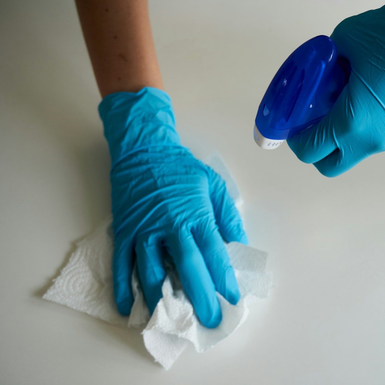 Gloved hands wiping a white surface with disinfectant and a paper towel